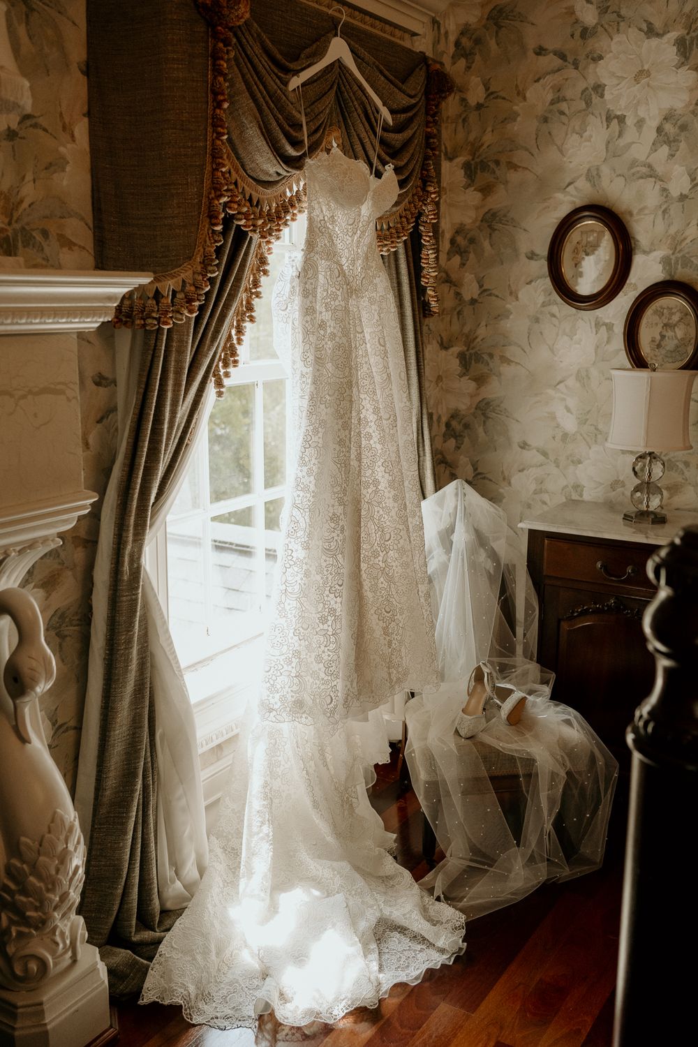Elegant lace wedding gown hanging by a window with ornate drapes in a vintage bridal suite.