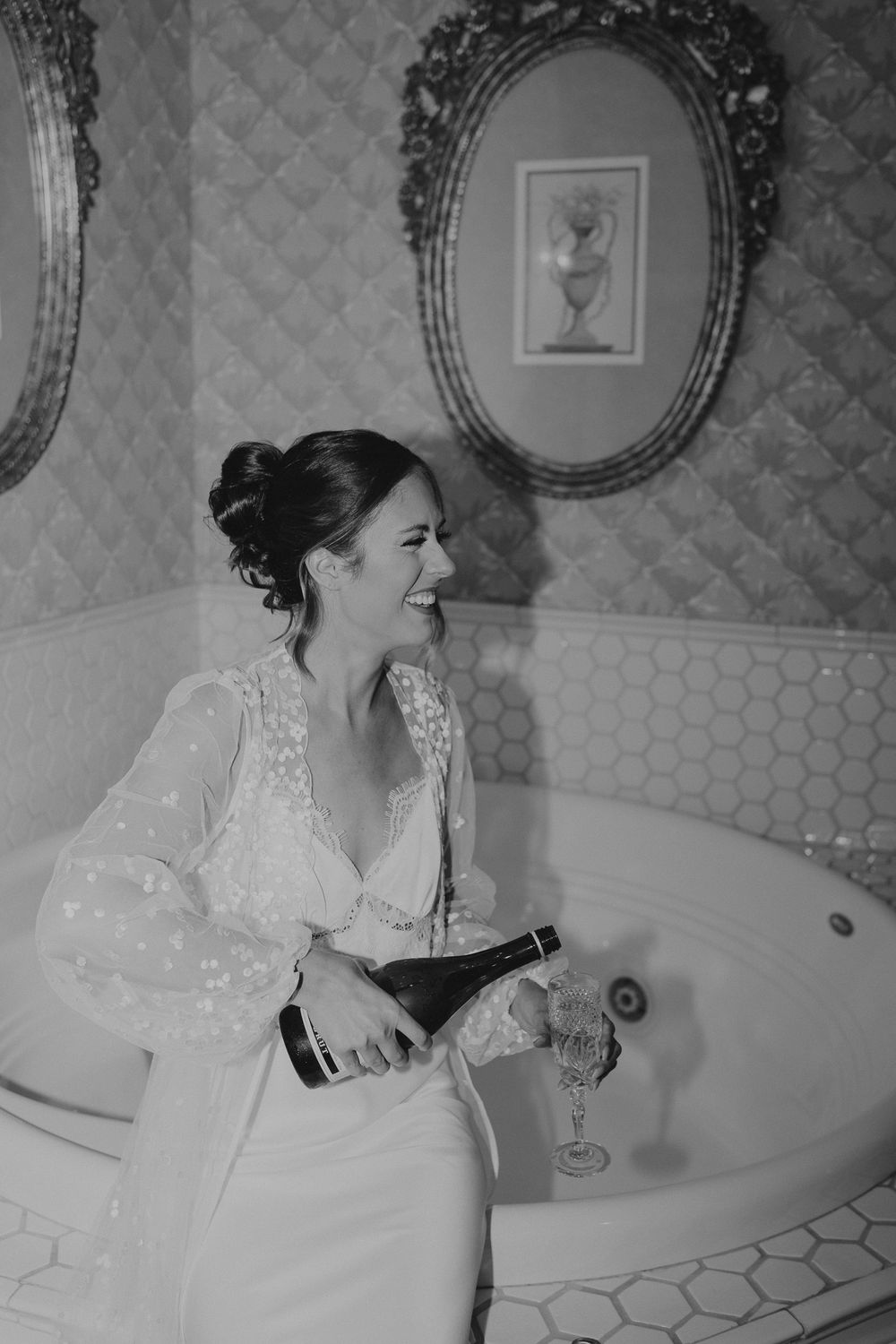 Joyful bride pouring champagne in vintage bathroom with ornate gold mirrors on wedding day.