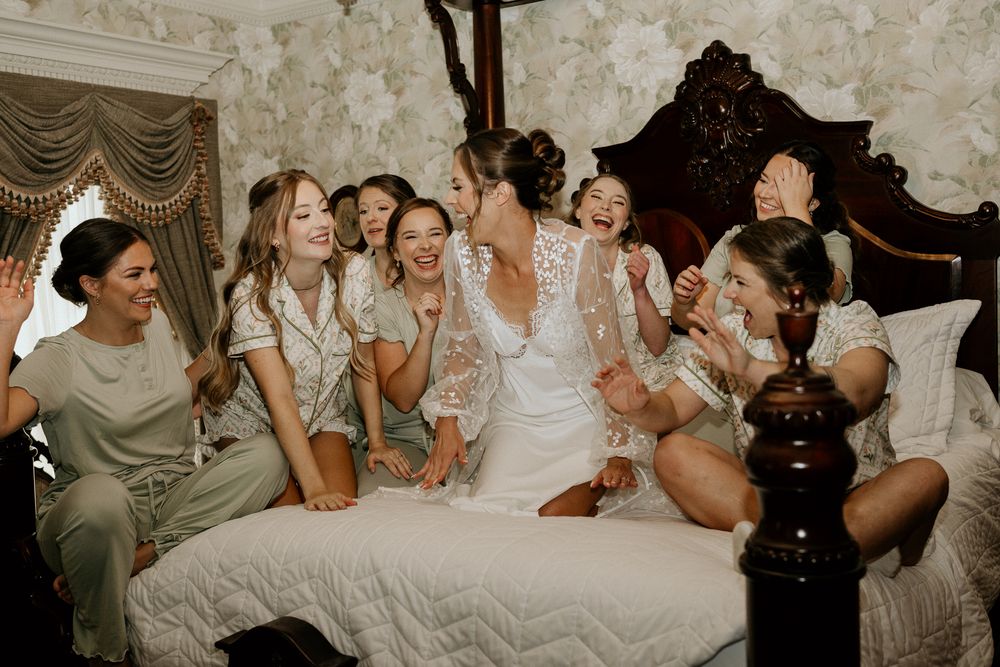 Joyful bride in lace gown celebrates with bridesmaids in sage satin robes on ornate bed in floral wallpapered room.
