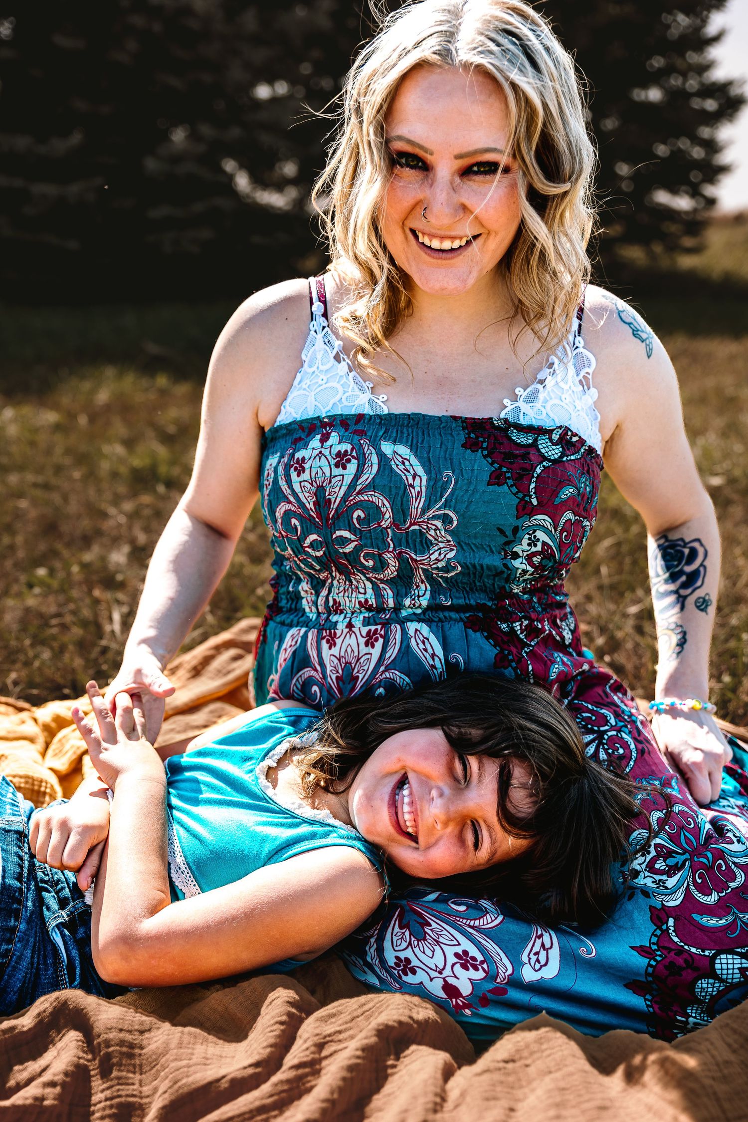 A laughing blonde woman in a floral dress sits outdoors while a smiling child lays across her lap on a blanket.