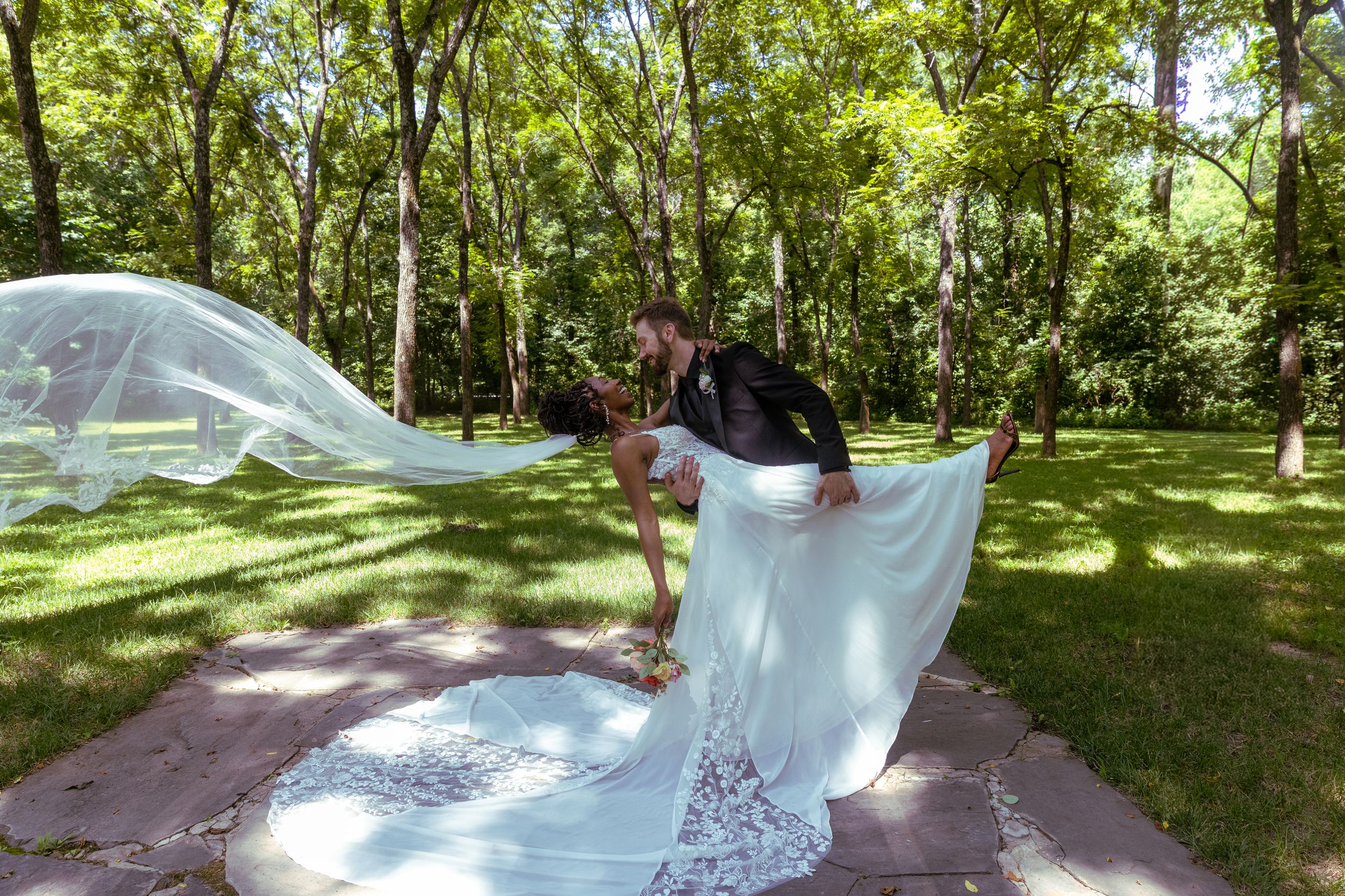 Groom dipping bride in white lace gown with flowing veil in a lush green park on their wedding day.
