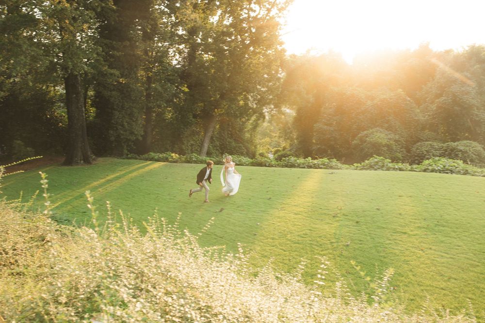 Bride and groom run joyfully across sunlit green lawn surrounded by lush trees.