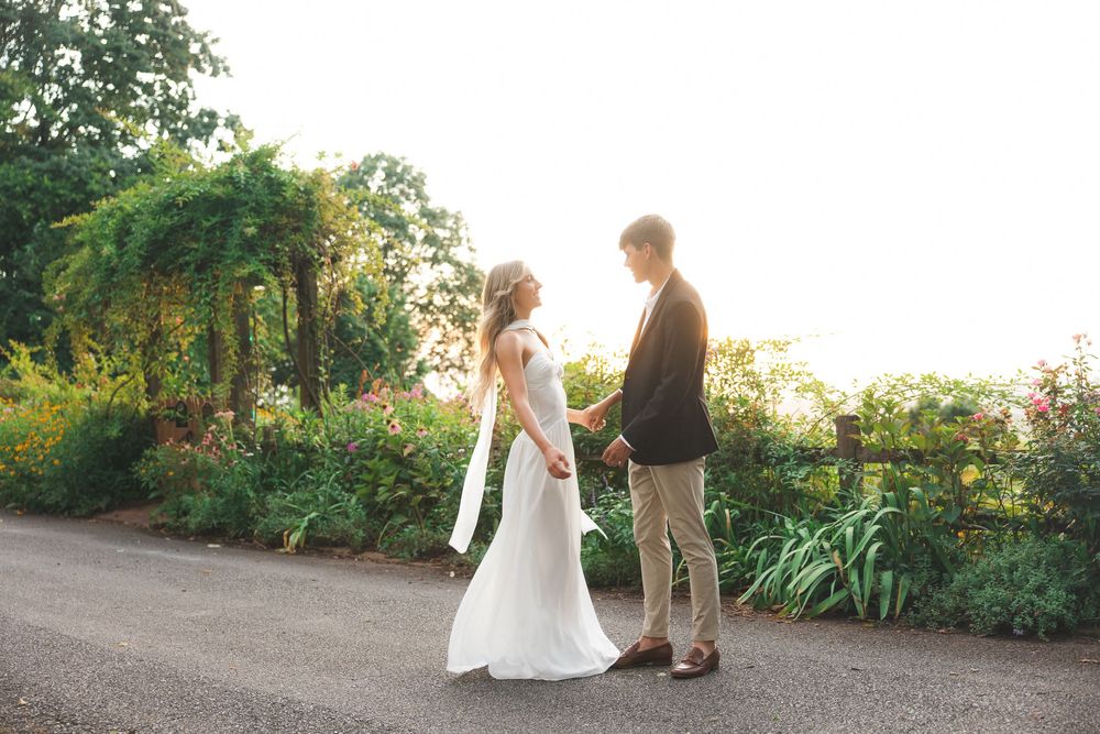 Bride and groom hold hands on garden path bathed in warm golden sunset.