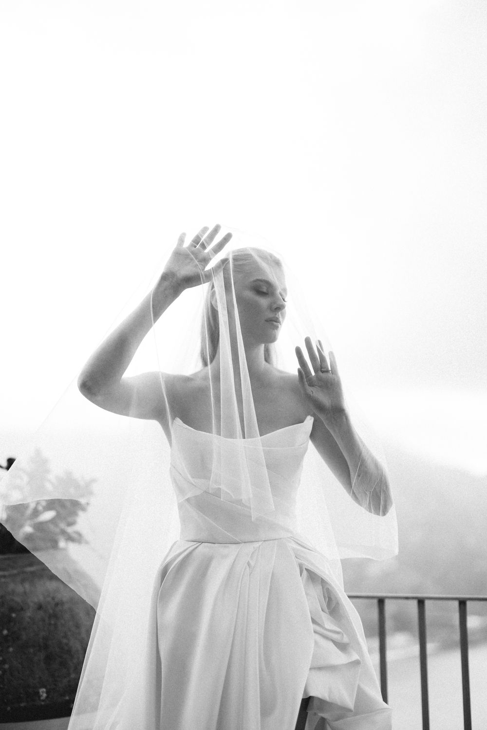 Black and white artistic portrait of bride in strapless gown lifting veil with both hands outdoors.