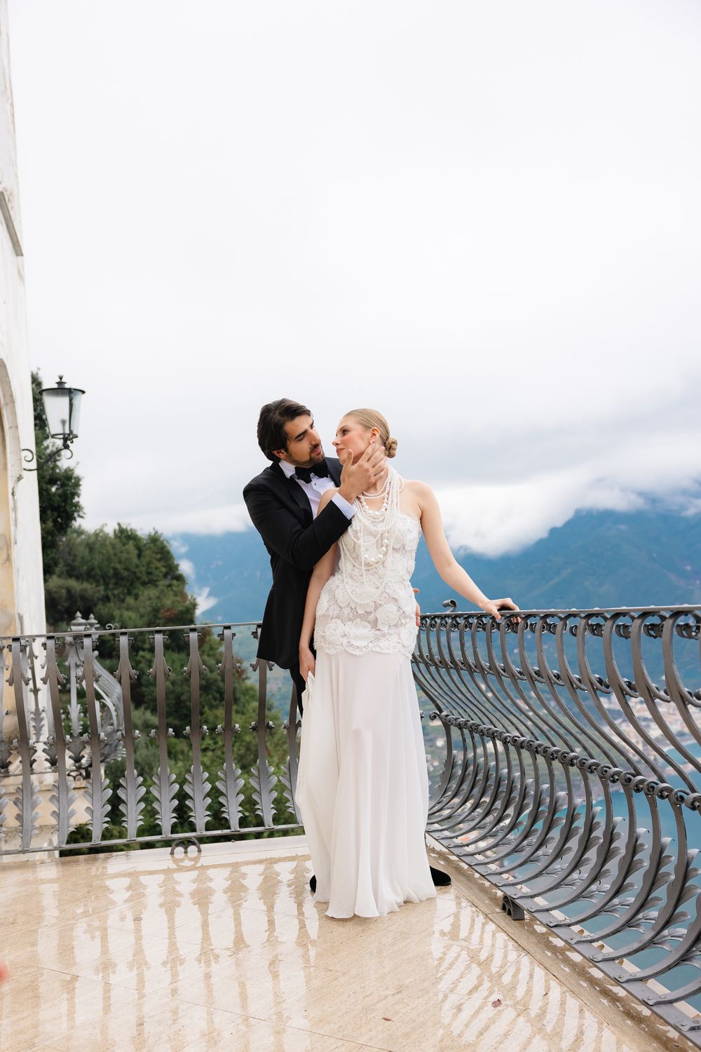 Groom in tuxedo embracing blonde bride in beaded halter gown on elegant balcony with mountain views.