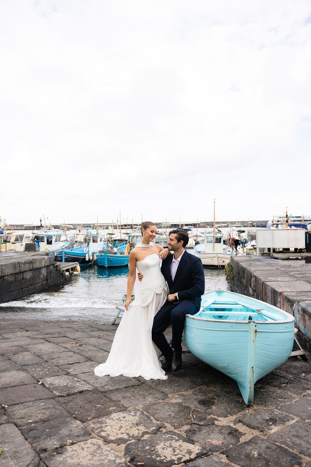 Bride and groom posing beside turquoise boat at scenic Italian harbor with boats in background.