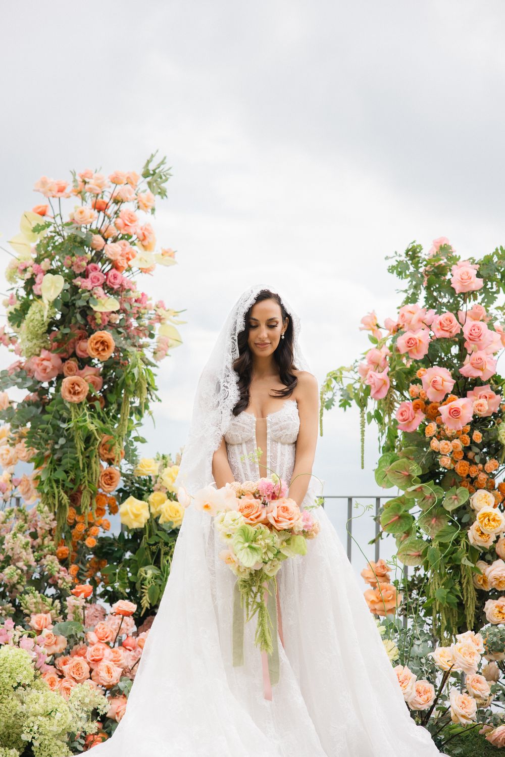 Bride in white corset gown holding peach bouquet surrounded by lush floral arrangements of roses in pink, orange, and yellow.