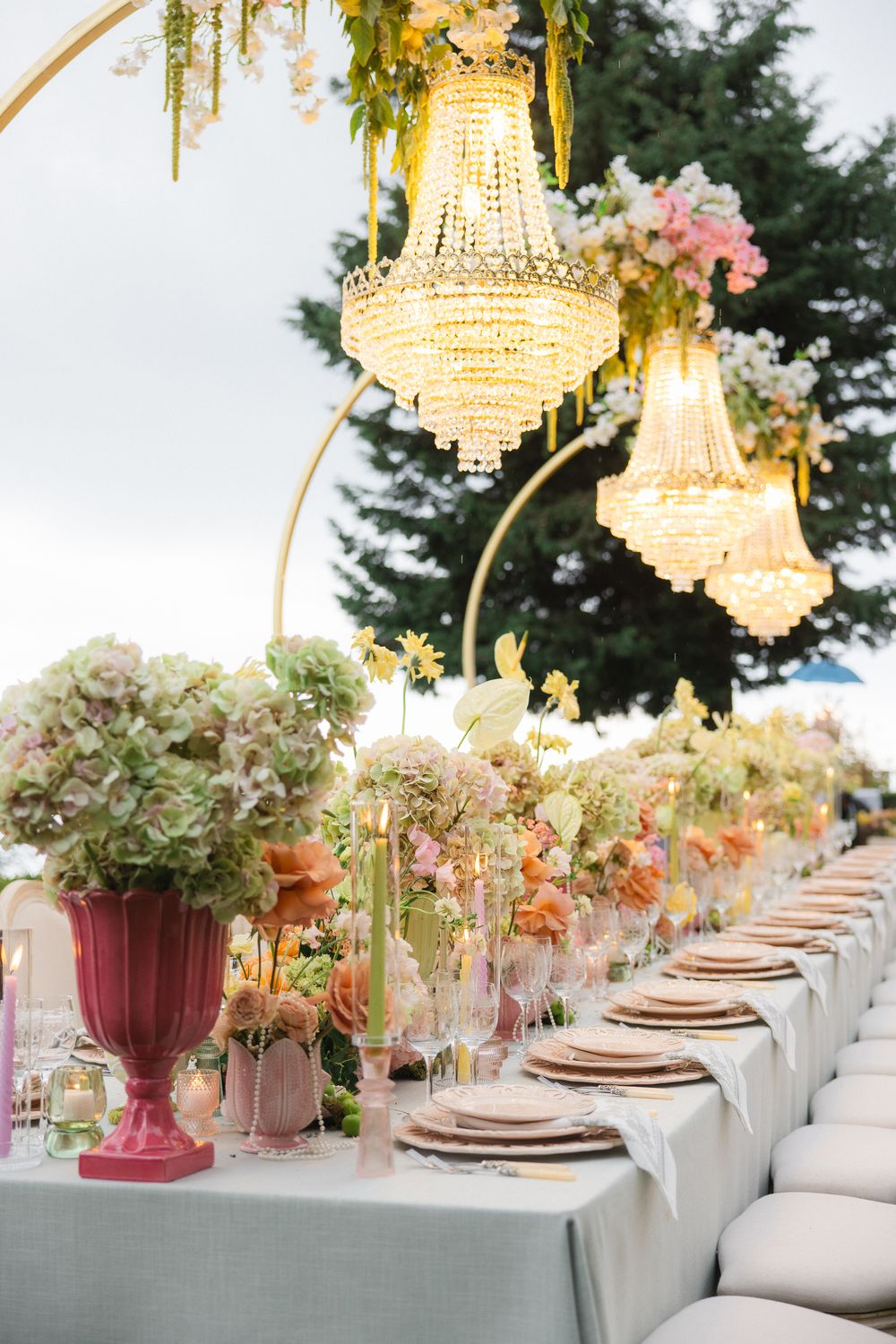 Elegant outdoor reception table with crystal chandeliers, hydrangea centerpieces, and gold settings.