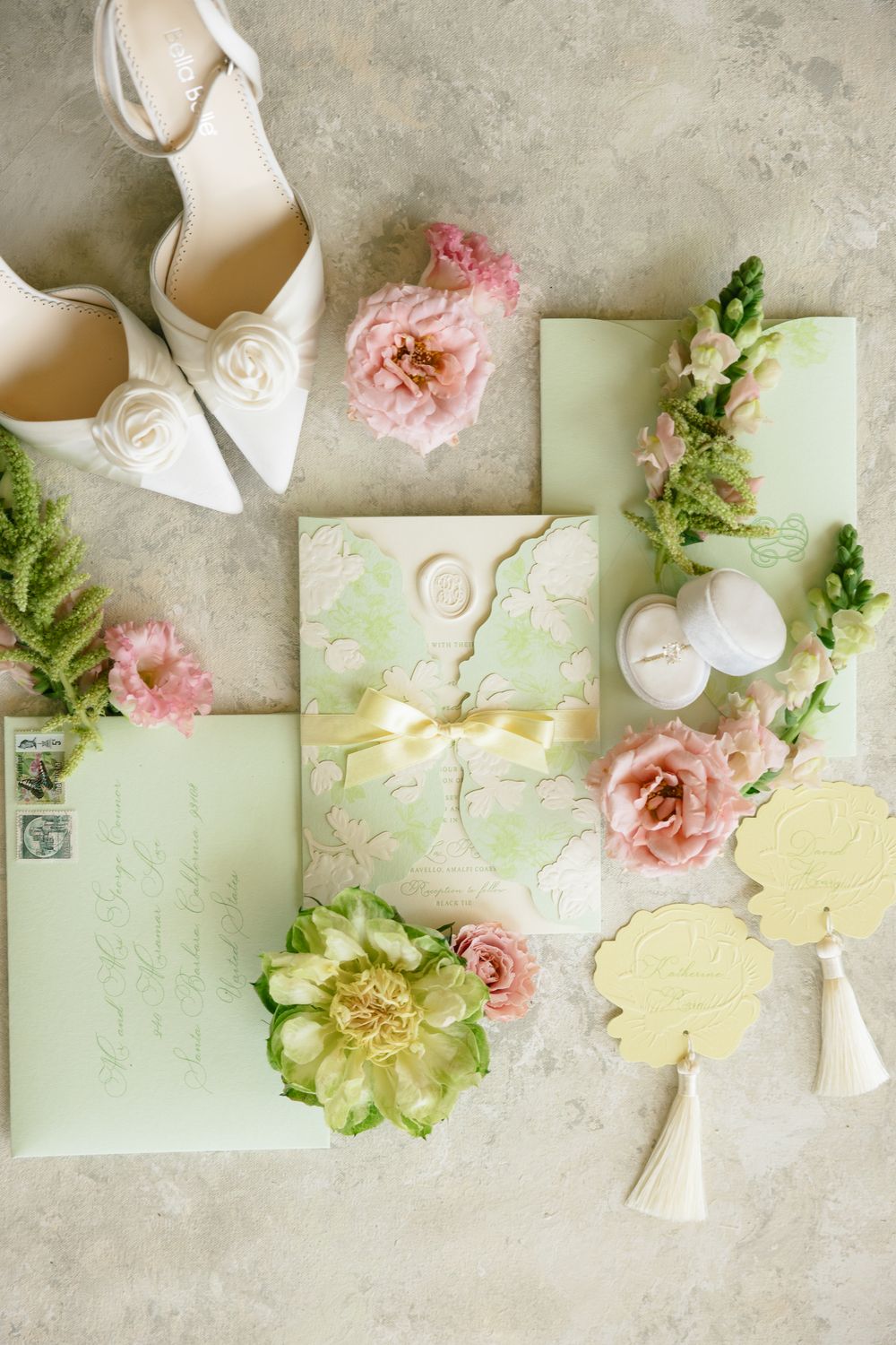 Flat lay of mint green wedding stationery, white heels, pink flowers, and gold earrings on marble.