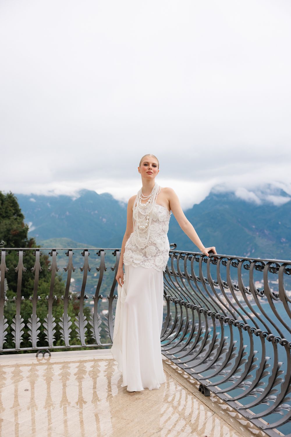 Elegant bride in white halter beaded gown standing on ornate balcony with misty mountain backdrop.