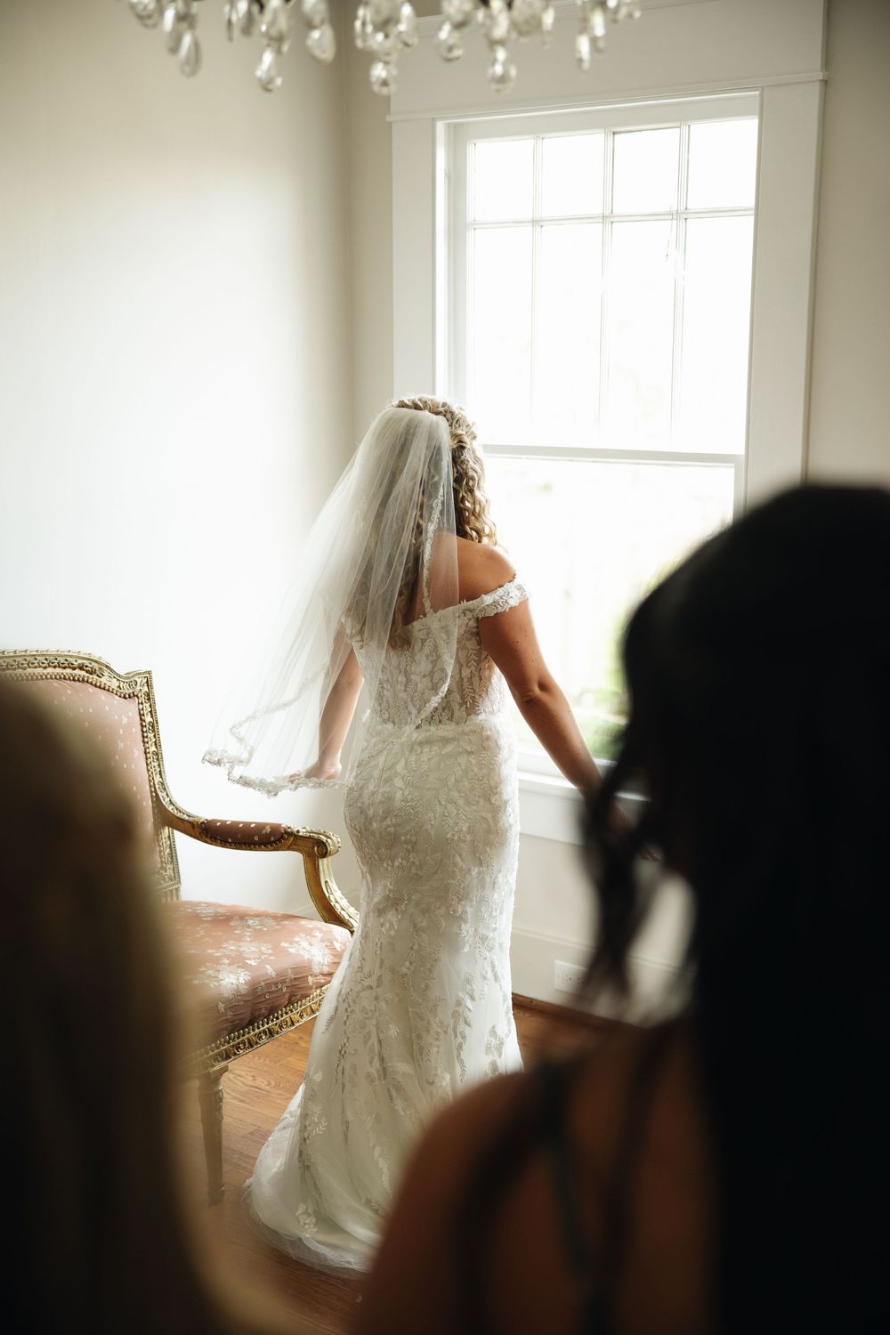 Bride in fitted lace wedding gown and veil stands by window while bridesmaids look on.