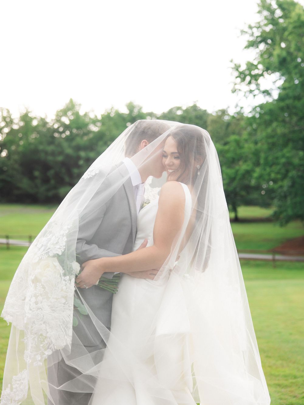 Smiling bride and groom sharing a romantic moment under a lace-trimmed veil on a green lawn.