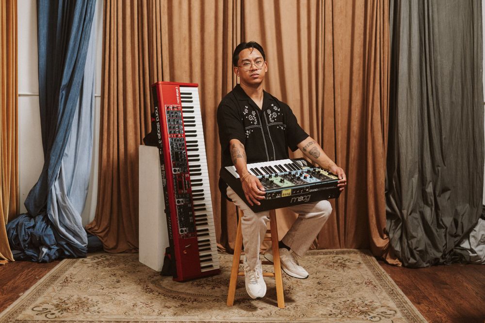 Musician poses with synthesizer and red keyboard against brown curtain backdrop in studio setting.