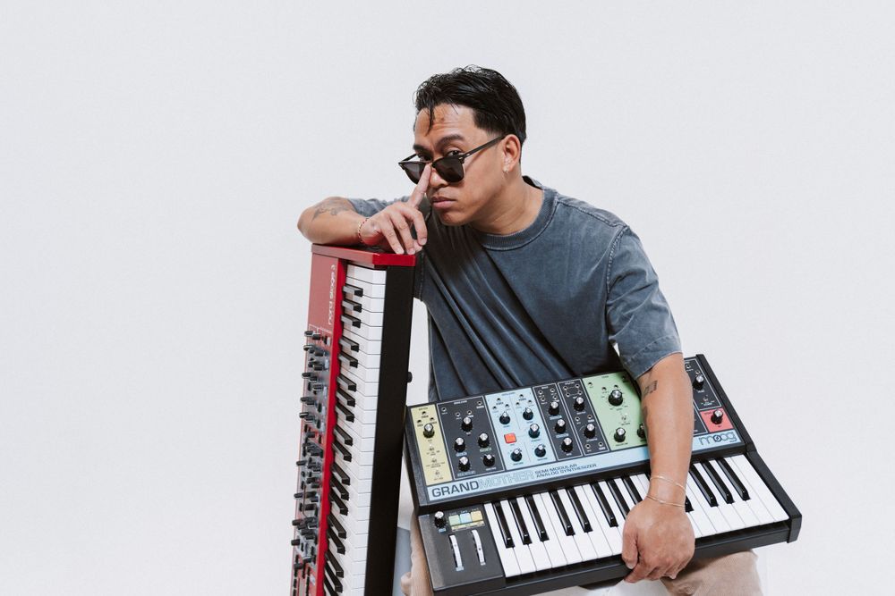 Musician posing with vintage synthesizer equipment against white background.