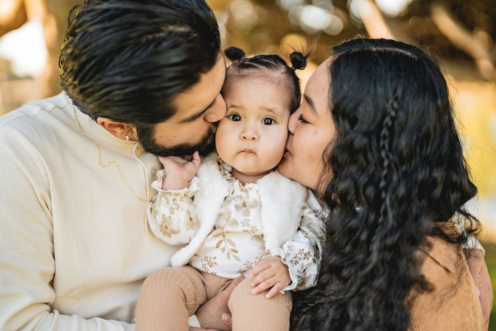Family sharing tender moment with baby in white dress outdoors.