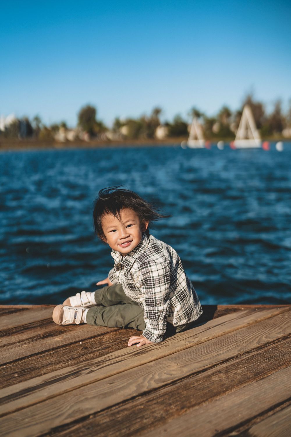Child in plaid shirt sitting on wooden dock by lake with sailboats in background.