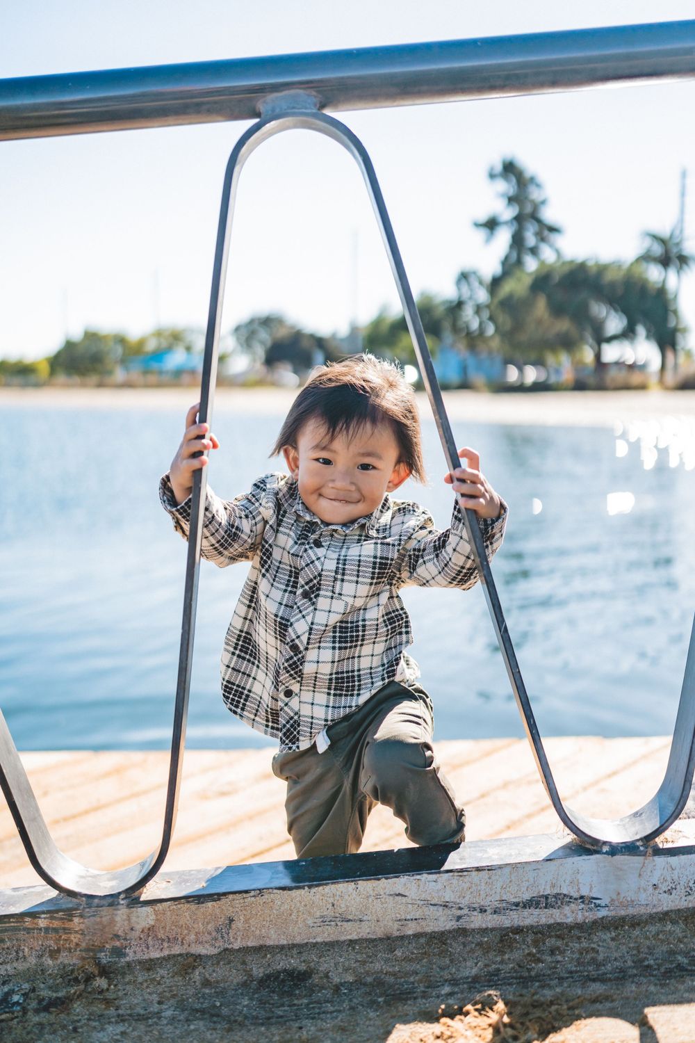 Child in plaid shirt playing on boat railing with water and palm trees in background.