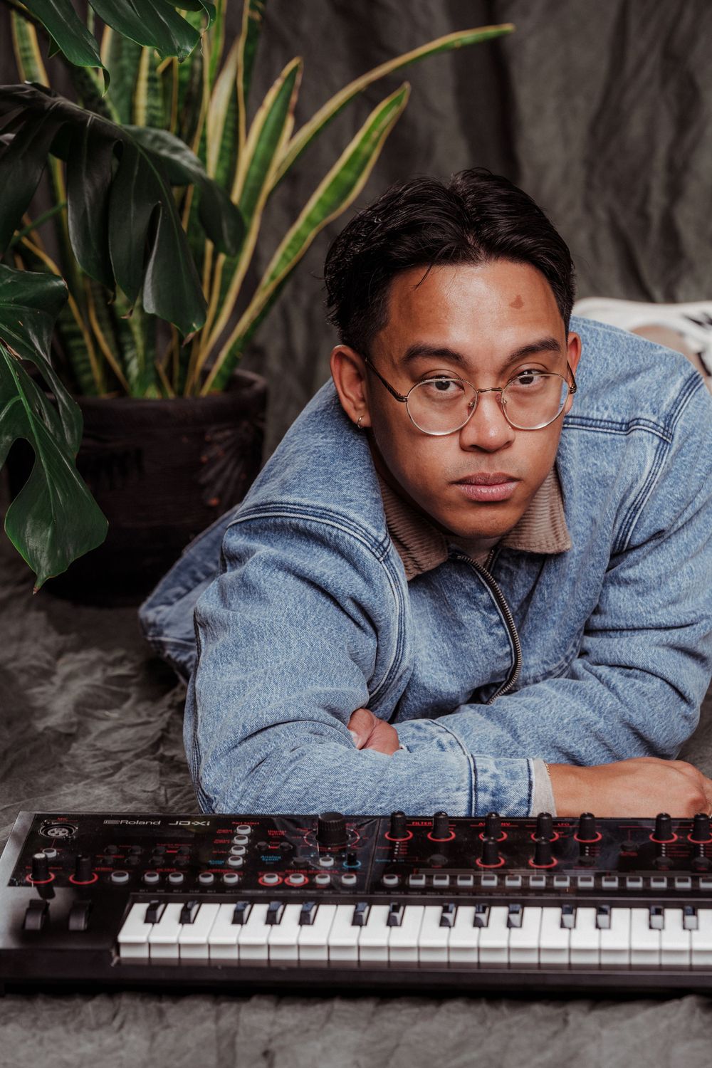 Artist with glasses composing music on electronic keyboard surrounded by plants.