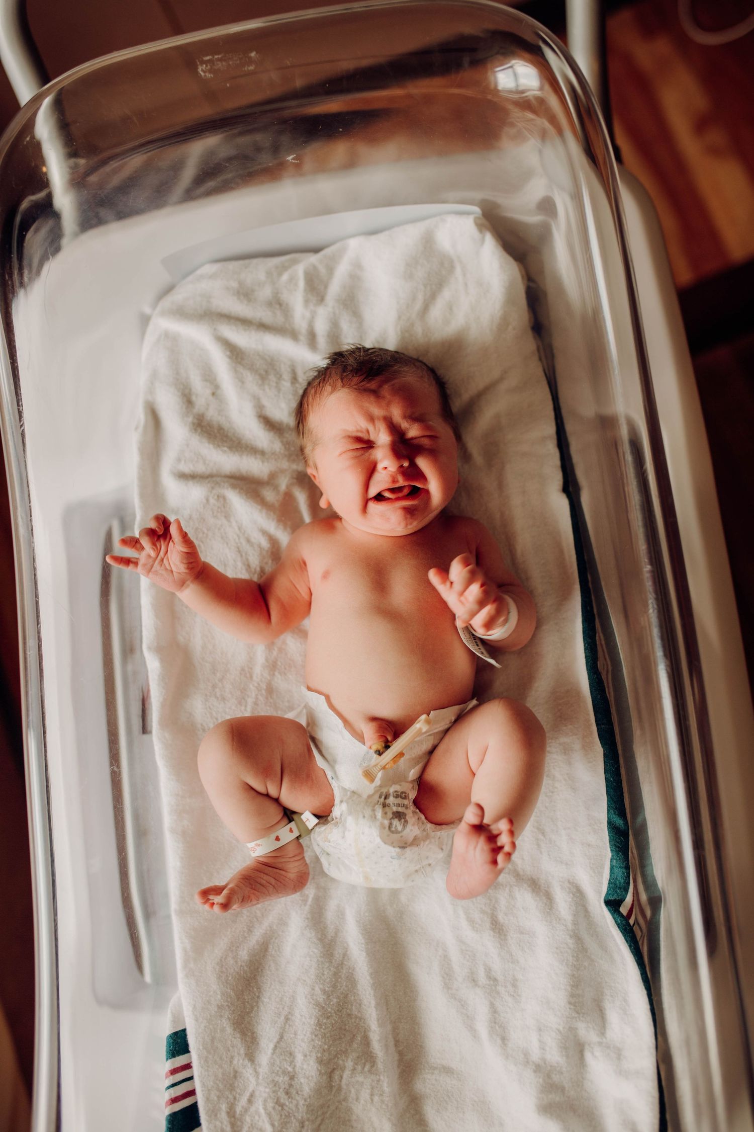 Newborn baby crying while lying in a hospital bassinet on white blanket.