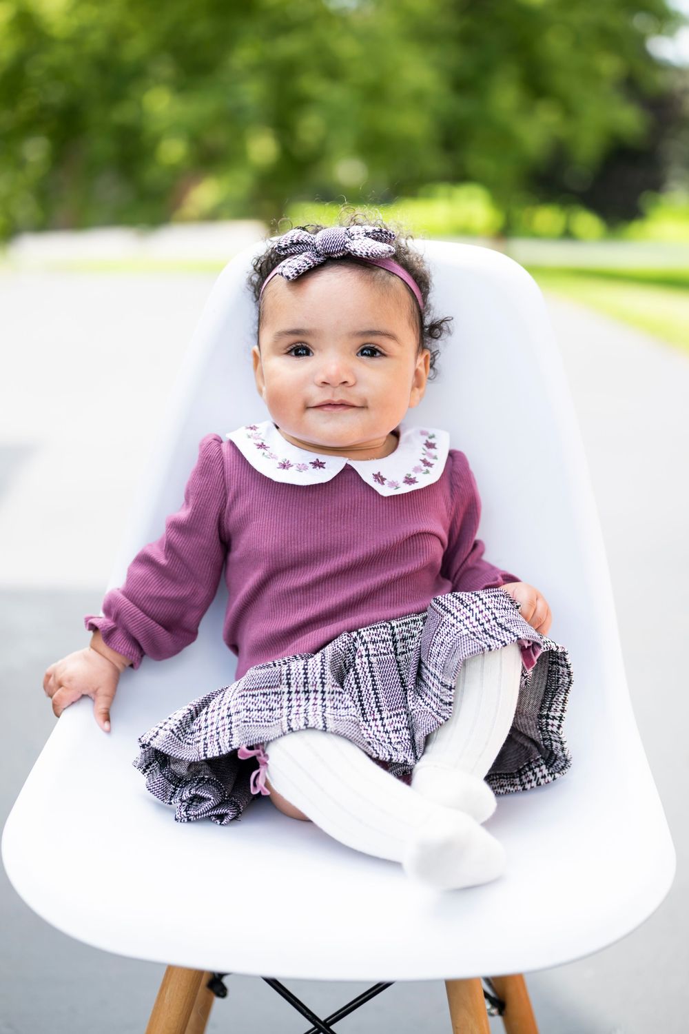 Adorable baby girl in purple top and plaid skirt sits in white modern chair outdoors, smiling at camera.