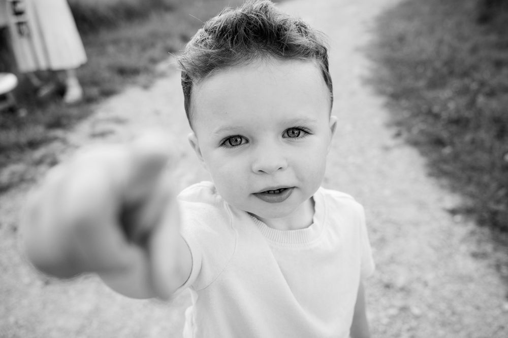 Black and white photo of a curious toddler boy reaching toward camera on a outdoor path.