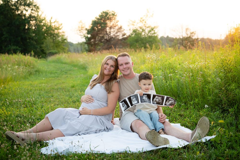 Pregnant woman sits with partner and toddler son holding ultrasound photos during outdoor maternity photoshoot in green field.