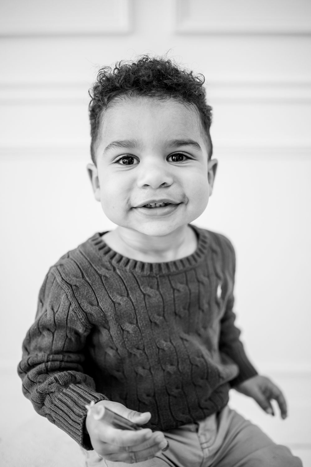 Black and white portrait of a smiling toddler boy with curly hair wearing a cable-knit sweater.