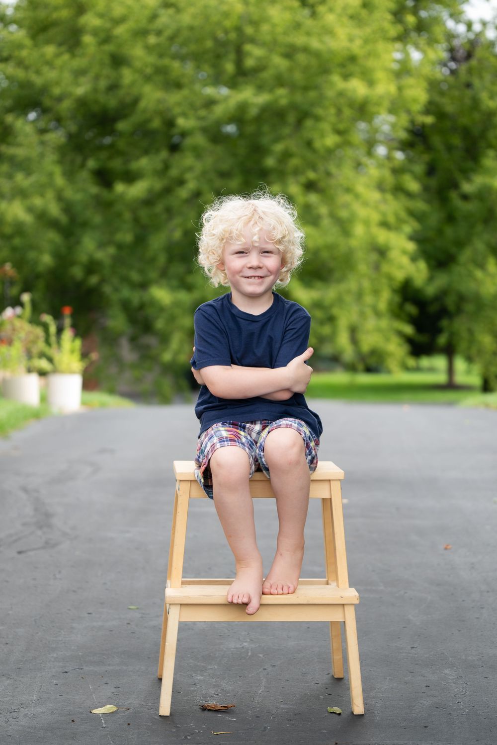 Smiling blond curly-haired boy sitting on wooden step stool outdoors with arms crossed, wearing navy shirt and plaid shorts.