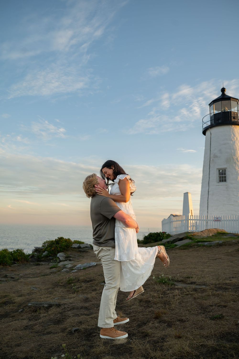 Couple embracing at sunset near a white lighthouse, man lifting woman in white dress by the ocean.
