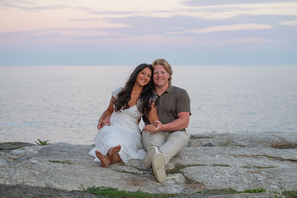 Smiling couple sitting close together on rocky oceanside cliff during pastel sunset.