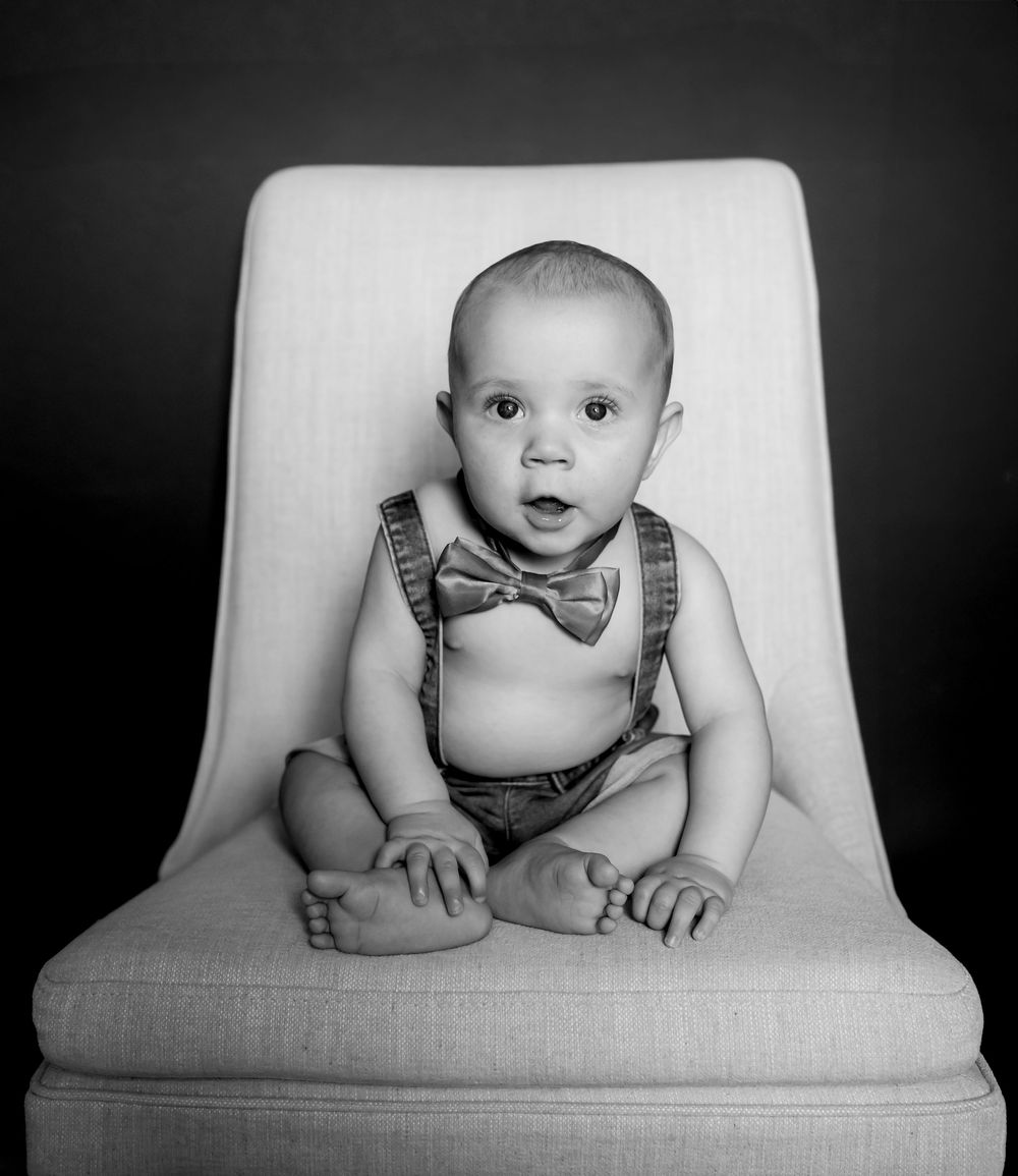 Adorable baby in bow tie and suspenders sitting in white chair, black and white portrait photo.