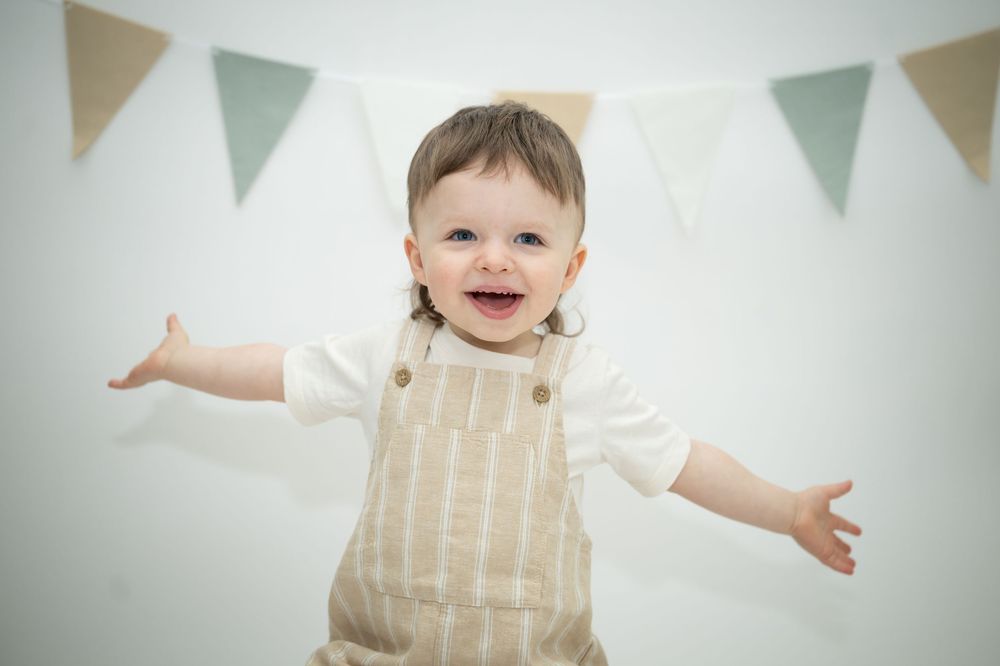 Happy baby in striped overalls smiling with arms outstretched, standing before a white wall with bunting flags.