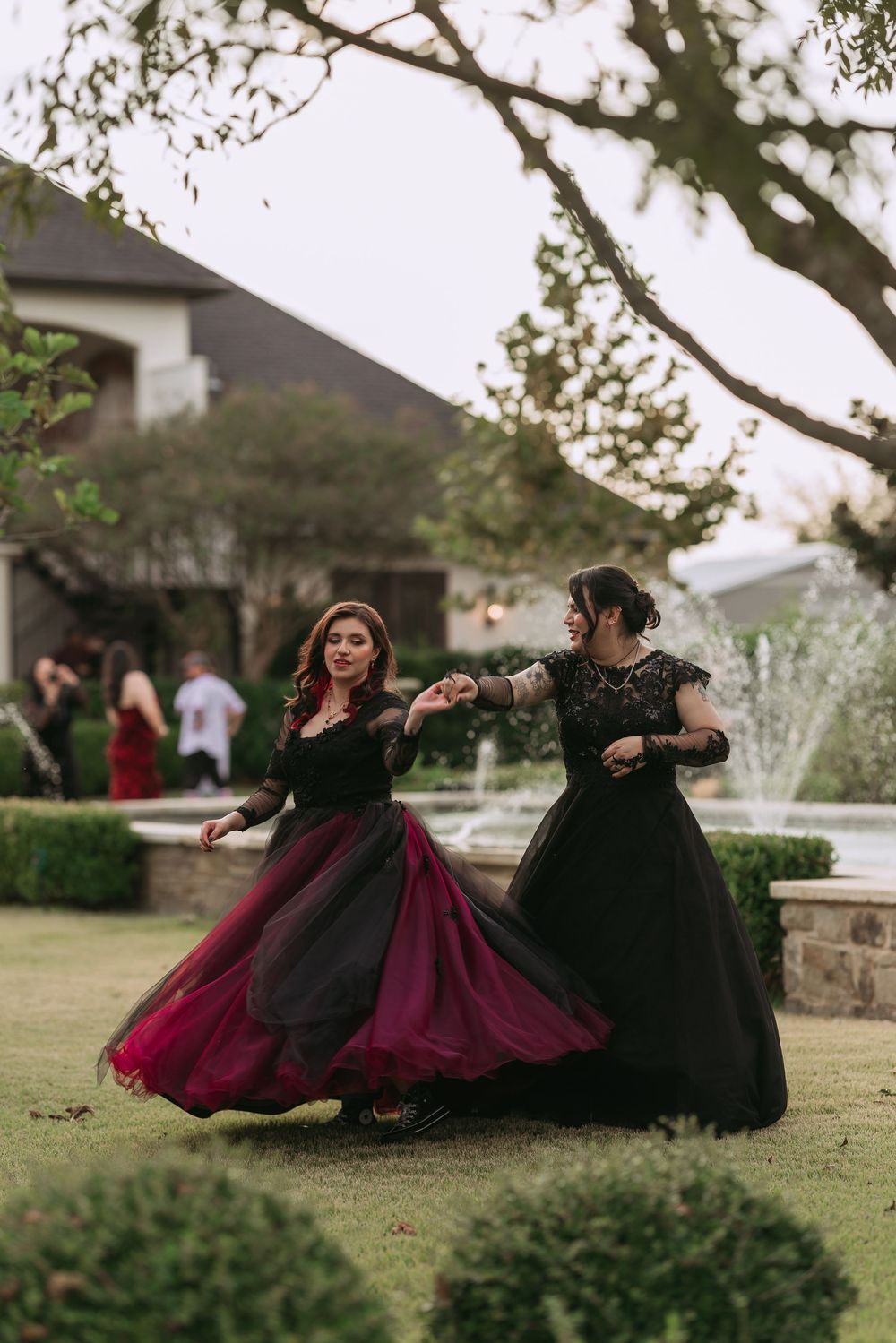 Two figures in black and burgundy dresses twirl and dance together on a grassy lawn near a residential building.