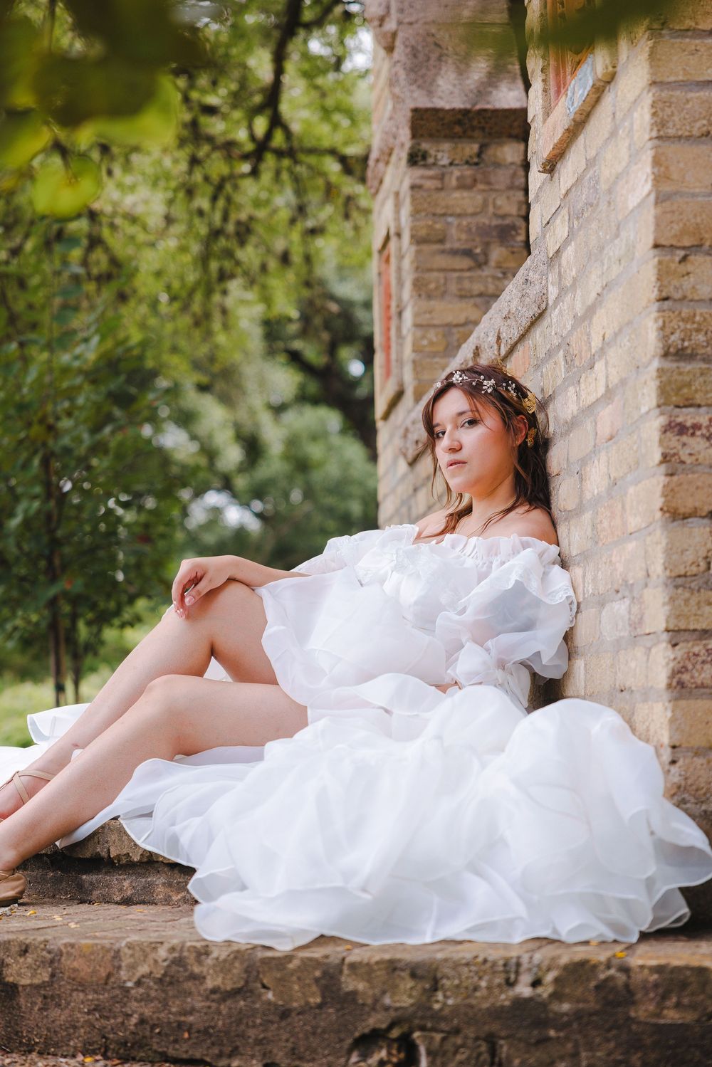 A bride in a flowing white wedding gown reclines elegantly against stone architecture in a garden setting.