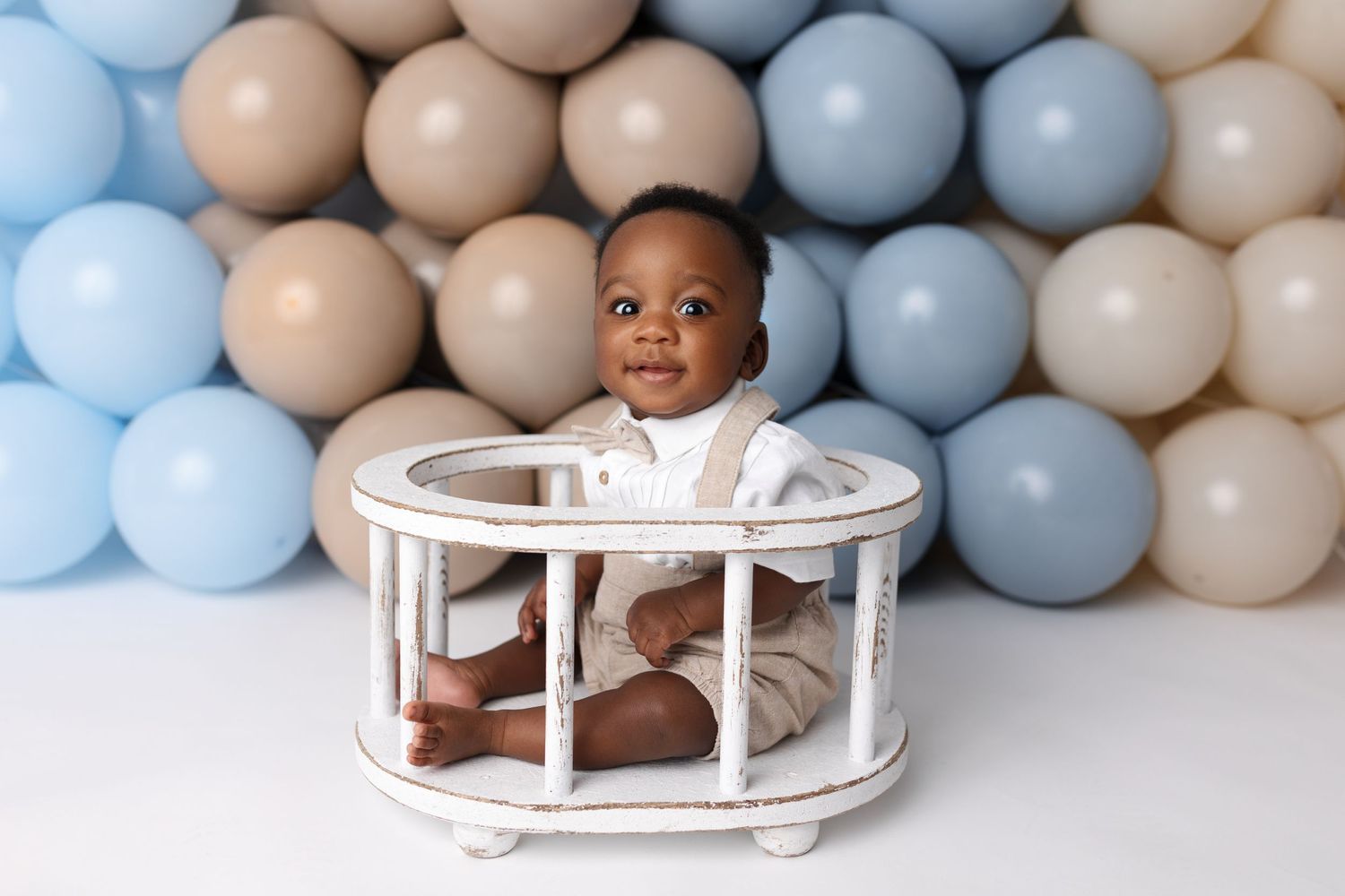 A baby poses in a white round wooden prop against a backdrop of blue and beige balloons for a portrait session.