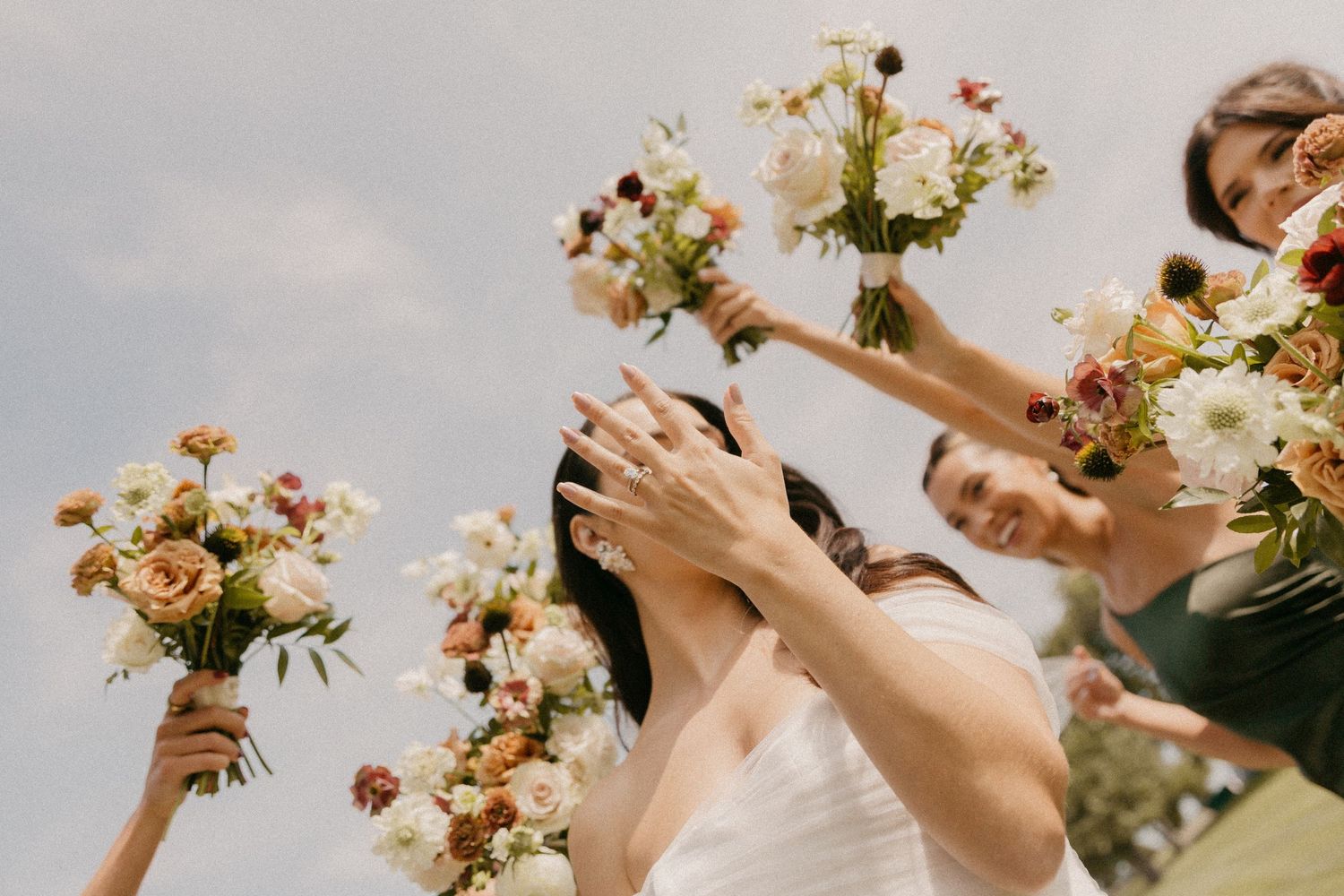 Wedding celebration scene with raised flower bouquets of cream and blush roses against a cloudy sky.