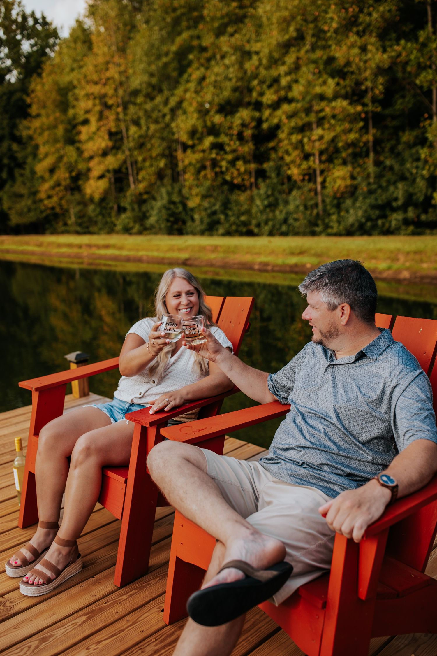 Couple relaxes in bright orange Adirondack chairs on wooden deck by lake.
