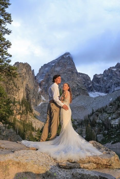 A couple in wedding attire stands on rocky terrain in the tetons with majestic mountain peaks and evergreen trees in the backdrop.