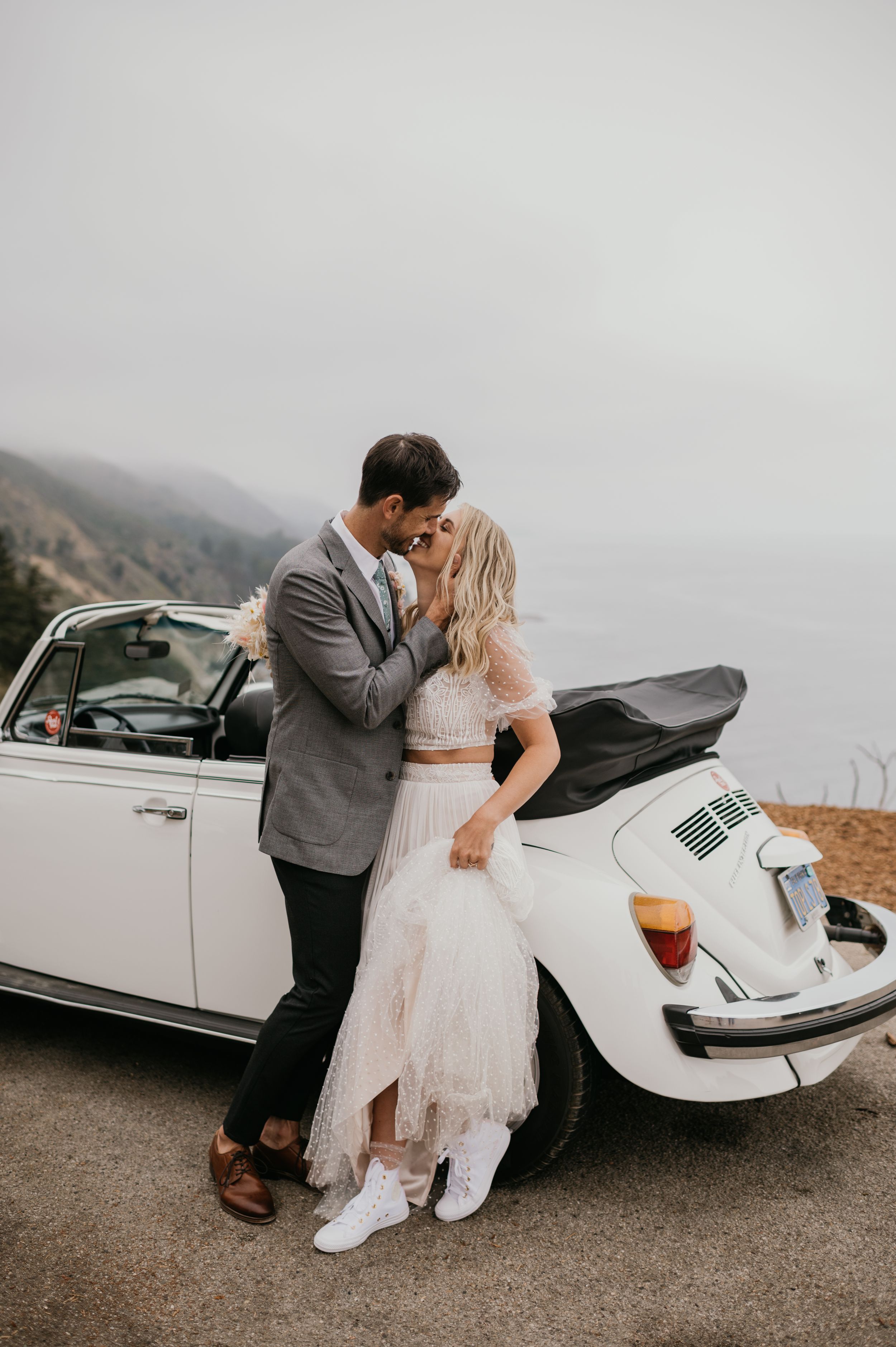 A couple in wedding attire poses romantically against a classic white Volkswagen Beetle on a misty road.