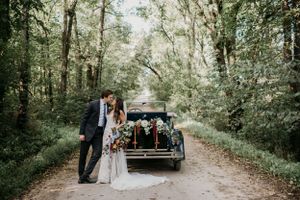 Newlyweds pose with vintage car on tree-lined path after their wedding ceremony.
