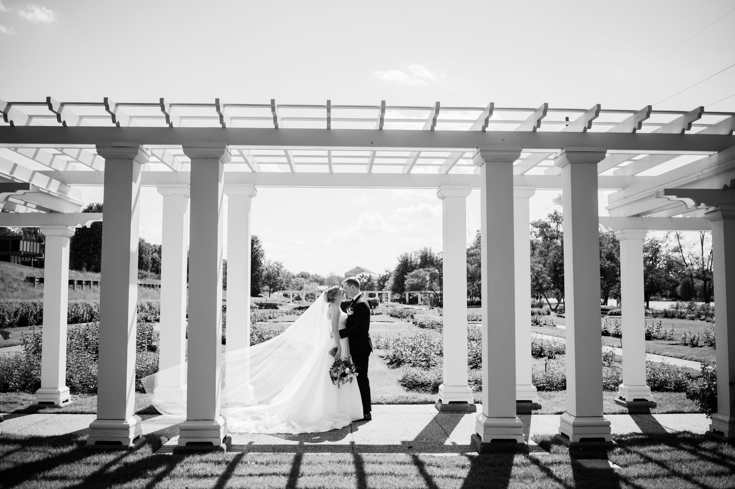 Bride and groom share a romantic moment under a white pergola in a beautiful garden setting.