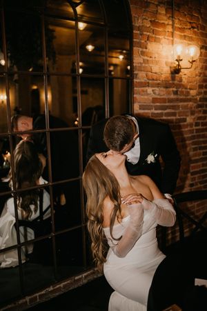 Bride and groom share a romantic dip kiss in front of a mirror at a warmly lit brick-walled wedding venue.