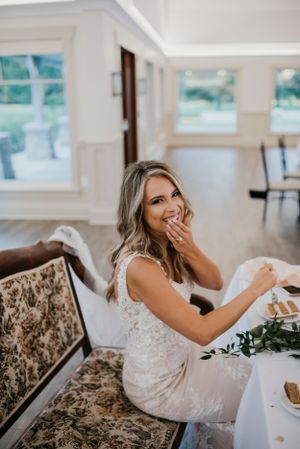 A person sits on an ornate vintage sofa while wearing a white lace wedding dress in a bright interior setting.