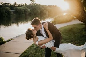 Groom dips bride for a romantic kiss at golden hour by a serene lakeside during their outdoor wedding ceremony.