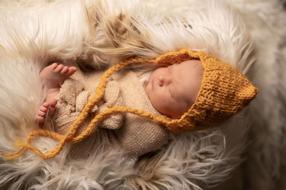 A newborn baby wearing a knitted yellow bonnet sleeps peacefully on soft white fur.