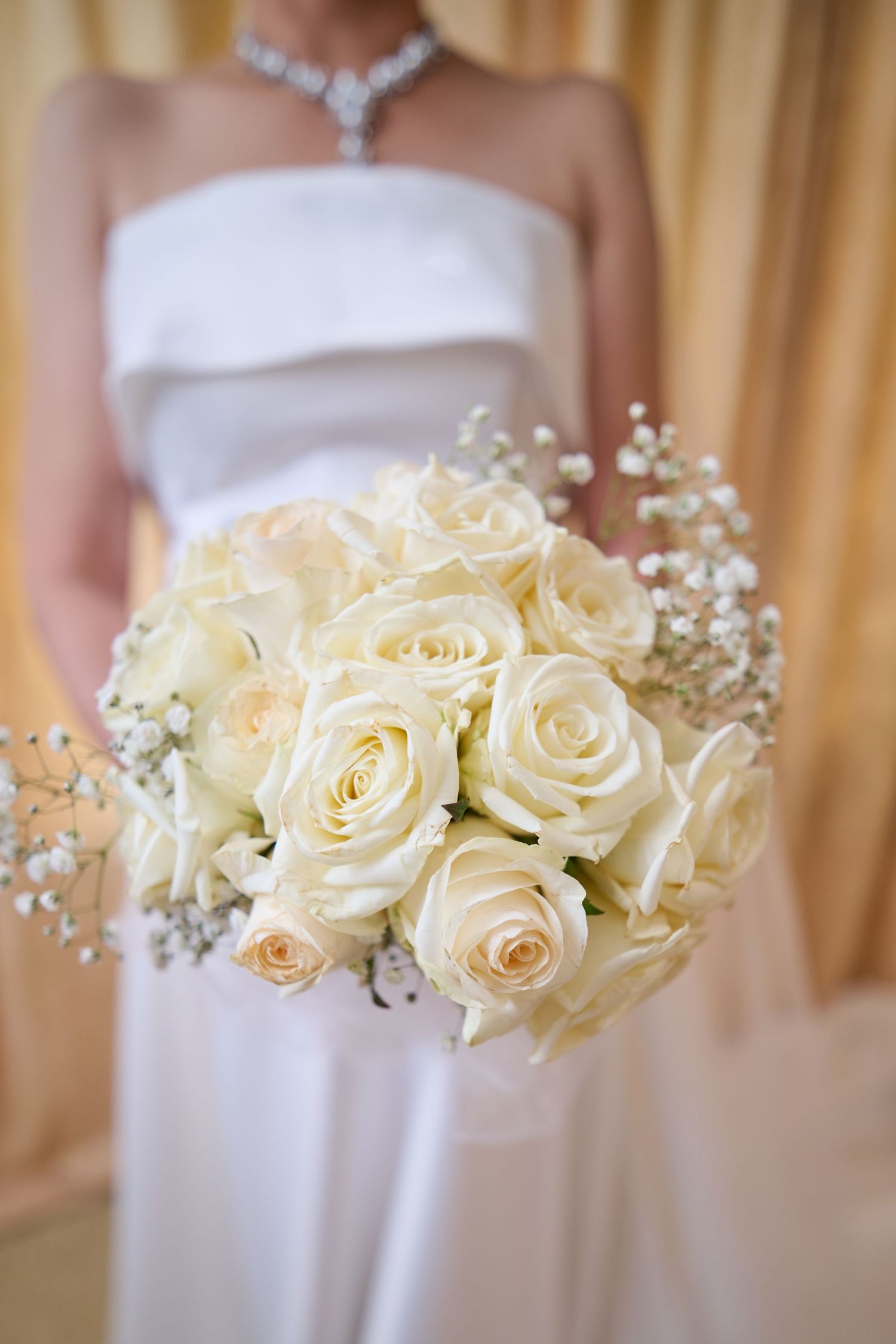 A delicate bridal bouquet of cream-colored roses and baby's breath held against a white dress with a sparkling necklace.