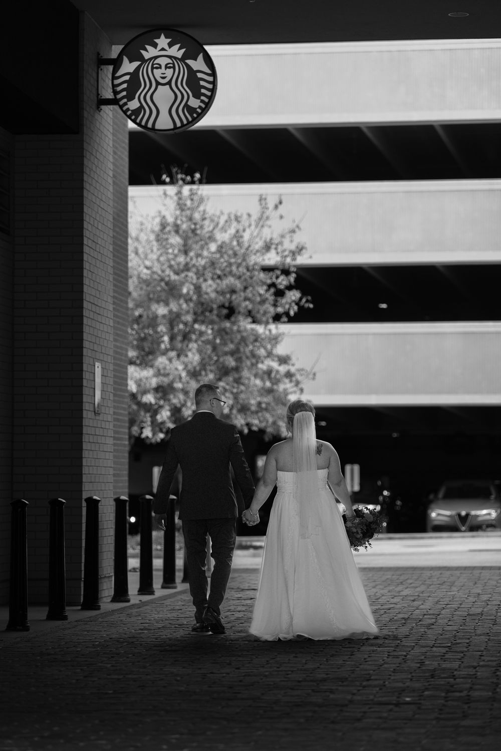 Bride and groom walk away from camera holding hands and coffee near a starbucks
