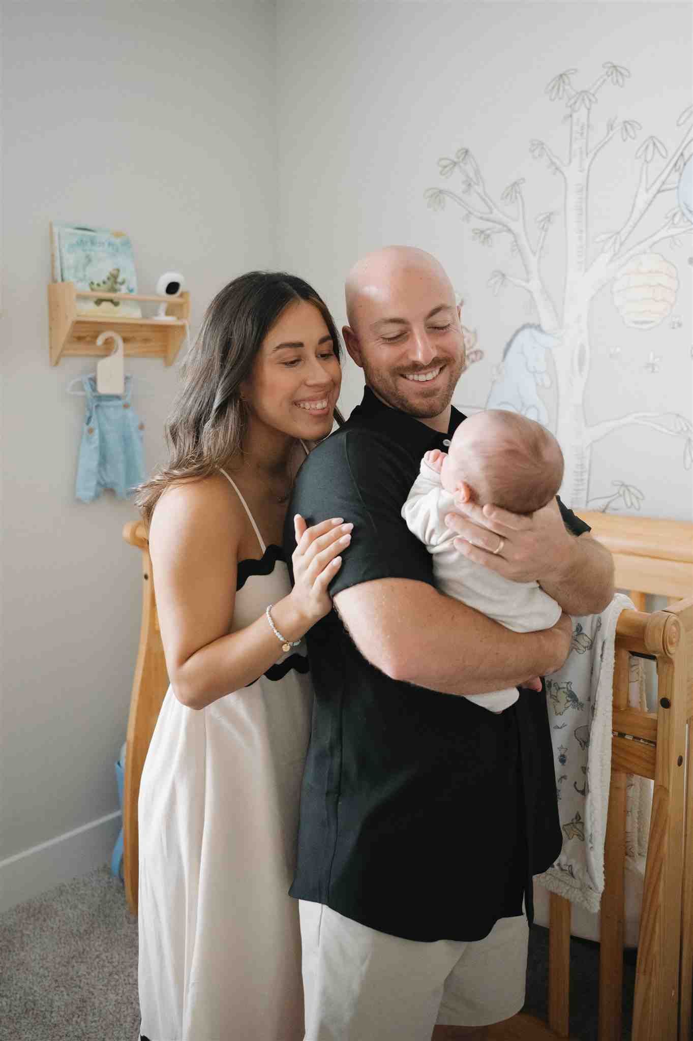 A couple shares a tender moment with their newborn baby in a nursery decorated with wall art and baby clothes.