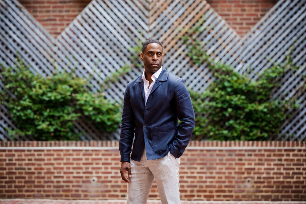 Professional portrait of a male in a navy blazer and light pants standing in front of a lattice and brick wall, by Justin Baker Photography.