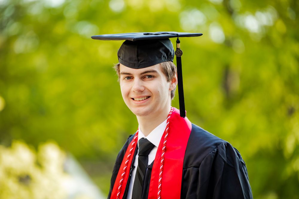 Graduate in cap and gown with red graduation stole poses outdoors with green trees in background in Maryland, by Justin Baker Photography.
